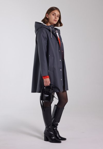 Grå vandtæt jakke med hætte, trykknapper og sidelommer, parret med knæhøje blanke sorte støvler og en lille sort håndtaske.