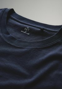 Sötétkék pamut póló kerek nyakú kivitelben. A belső nyakrészen nyomtatott felirattal rendelkezik: "THE SET M" és "MADE IN BANGLADESH". Puha tapintású.