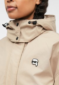 Beige vízálló dzseki magas gallérral, patent gombokkal és fekete rajzfilmes logóval a mellkason. Simább textúra, állítható húzózsinórral.