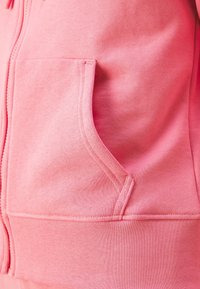 Gros plan sur la poche d'un sweat à capuche zippé rose avec des coutures visibles et la texture du tissu.