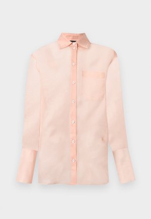 Kvinde stående barfodet iført en lyserød gennemsigtig langærmet skjorte med knapper og matchende shorts mod en ensfarvet hvid baggrund.