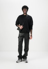 Sweat-shirt noir, pantalon cargo en cuir noir et baskets à motifs. Tient un petit sac noir avec une bandoulière. Design minimaliste.