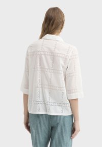 Chemise blanche légère avec un col orné de découpes œillets disposées en motif quadrillé. Manches trois-quarts et coupe décontractée.