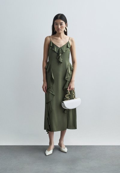 Kvind i en olivengrøn ruffle midi-kjole, der holder en hvid håndtaske med bølget guldhåndtag, iført hvide spidse flade sko og store guld hoop øreringe.