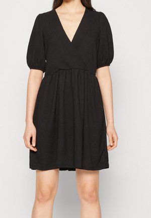 Femme portant une robe courte noire avec des manches courtes bouffantes et un décolleté en V profond, debout devant un fond clair uni.