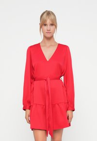 Robe rouge avec un décolleté en V, des manches longues, une ceinture à la taille et une jupe à volants. Fabriquée en tissu lisse avec un éclat subtil.