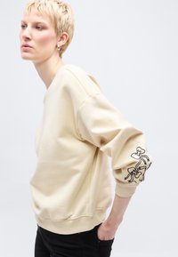 Άτομο με κοντά ξανθά μαλλιά φοράει μπεζ φούτερ με μαύρο κέντημα φιόγκου στο μανίκι και μαύρο παντελόνι, ποζάροντας μπροστά σε λευκό φόντο.