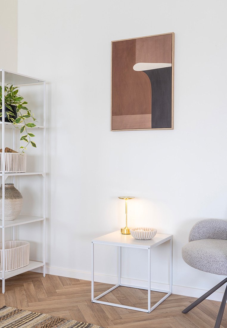 Minimalistische Ecke mit einem weißen Metallschrank, der Pflanzen und Körbe hält, einem kleinen weißen Tisch mit einer goldenen Lampe und einer Schüssel sowie einem geschwungenen grauen Stuhl.