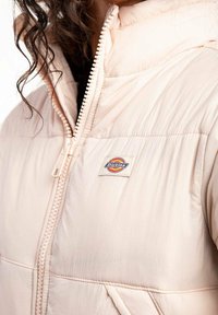 Chaqueta acolchada de color rosa claro con cremallera frontal, textura acanalada y un parche con el logo. Cuenta con capucha y bolsillos laterales.