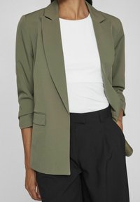 Blazer vert olive avec une coupe ajustée, fermeture à un bouton, et manches retroussées, associé à un haut blanc et un pantalon noir.