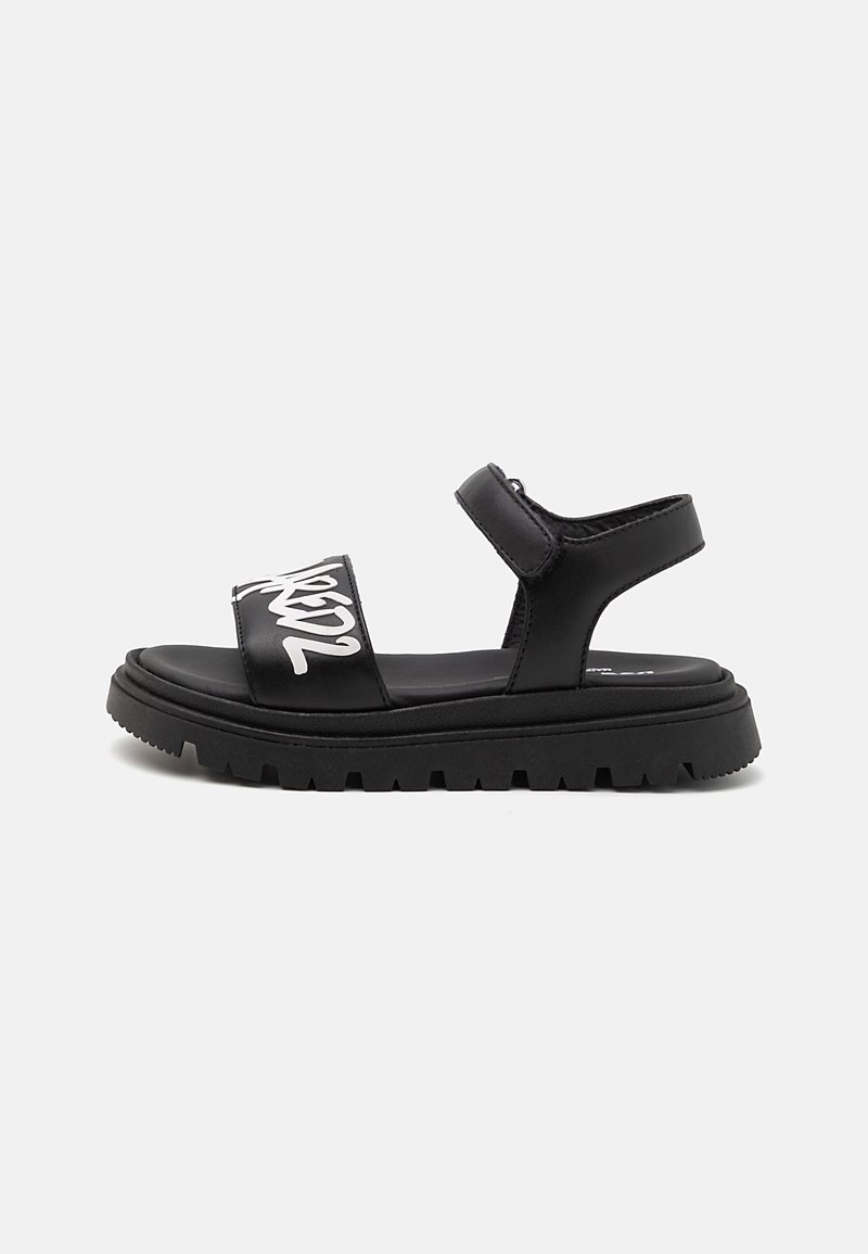 Black sandal with two wide straps, featuring white text. Thick sole with a textured, rugged bottom for grip. Adjustable ankle strap.