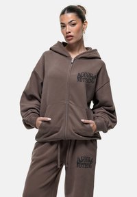 Felpa con zip di colore marrone, dotata di grandi tasche frontali e logo stampato in nero sul lato sinistro del petto. Tessuto morbido con vestibilità comoda e cappuccio.