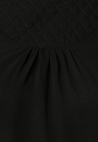 Svart blus med ett kviltat mönster vid halsen, med samlat tyg som faller mjukt. Mjukt material med en matta yta.