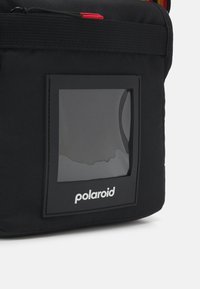 Sac en toile noir avec une fenêtre rectangulaire transparent et un logo "Polaroid" blanc ; comprend un tirette de fermeture éclair rouge et des détails de couture durables.
