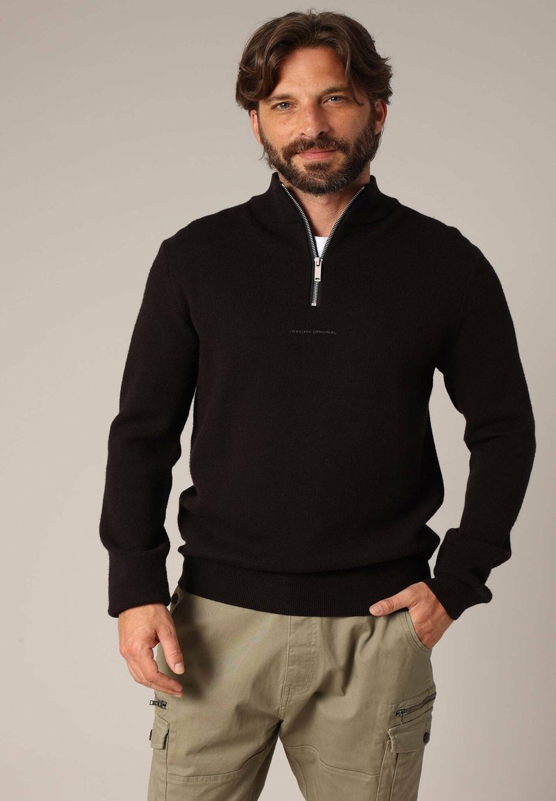 Zwarte ritstrui met lange mouwen en een hoge kraag, gemaakt van zacht materiaal. Draagt bij lichte cargo-broeken.