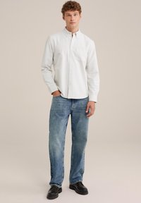 Chemise blanche à manches longues avec col boutonné, poche poitrine ; associée à un jean droit bleu clair et des chaussures noires.