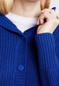 Niebieski dziergany kardigan o prążkowanej fakturze, z przodem na guziki i szerokim kołnierzem. Posiada okrągłe niebieskie guziki i średnią grubość.