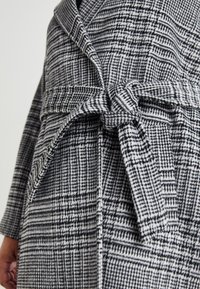 Μαύρο και άσπρο καρό παλτό με ζώνη. Διαθέτει γιακά σαλονιού, υφασμάτινη υφή και πλαϊνές τσέπες. Λεπτομέρεια κόμπου στη μέση.