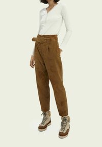 Vrouw in hoog-zittende bruine suède broek met een bijpassende riem, witte lange mouwen henley top en beige wandelschoenen met bontafwerking.