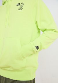 Neon grøn hættejakke med en frontlomme, sort Nike-logo på ærmet og et lille astronautgrafik på brystet. Materialet er i bomuld og har en afslappet pasform.
