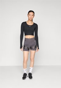 Crni dugih rukava crop top s pojasom "NIKE PRO", uparen s ljubičim prilagođenim kratkim hlačama s "NIKE PRO" uzorkom. Crne tenisice i bijele čarape.