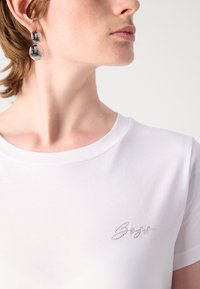 T-shirt bianco in cotone con scollatura rotonda e logo decorato con strass argentati sul lato sinistro. Indossato con orecchini pendenti metallici.
