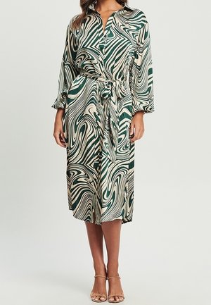 Kobieta ubrana w midi sukienkę koszulową w zielono-kremowe abstrakcyjne wzory z wiązaniem w talii i nude sandałki na obcasie.