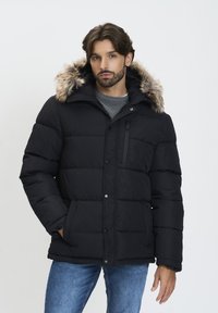 Chaqueta de plumas negra con capucha forrada de piel, presenta un bolsillo con cremallera y botones a presión. El material parece suave e insulated, con costuras horizontales.