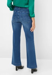 Wijde, blauwe denim jeans met een gladde textuur, vijf zakken en een middelhoge tailleband, gedragen met zwarte hakken en een groene bloes.
