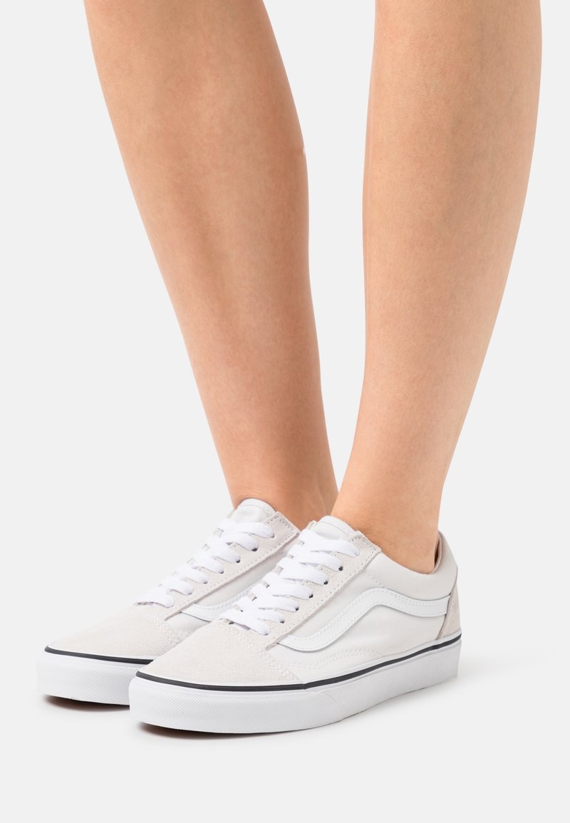 Personne portant des baskets blanches basses à lacets avec une bande noire sur la semelle blanche, debout sur un fond clair uni.