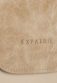 Beige läderhandväska med en texturerad yta, med präglar "EXPATRIÉ"-logotyp. Släta kanter och minimalistisk design.