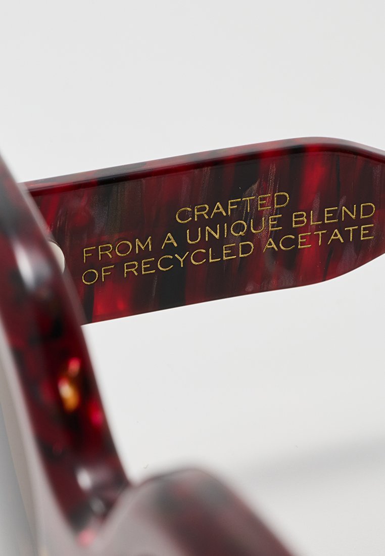 Rot-braun marmorierter Acetat-Brillenrahmen mit golden eingraviertem Text an dem Bügel, der lautet: "Hergestellt aus einer einzigartigen Mischung aus recyceltem Acetat."