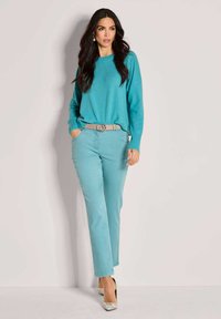 Frau mit langen dunklen Haaren, die einen türkisfarbenen Pullover, passende türkisfarbene Hosen mit beigem Gürtel und gemusterte High Heels trägt, steht vor einem einfarbigen Hintergrund.