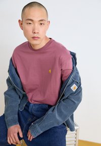 Denim jacket over a short-sleeve maroon T-shirt, featuring a small logo. Blue jeans complete the outfit. Neutral background.
