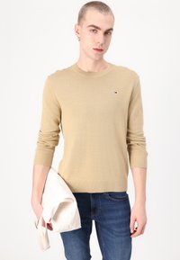 Beige strikket genser med ribbestrikkede mansjetter og kant, som har en liten flerfarget logo på brystet. Modellen holder en lysfarget veske.