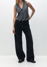 Grå ærmeløs vest med en dyb V-udskæring, knaplukning foran og to sidelommer, parret med brede sorte denimjeans og sorte flade sko.