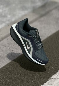 Ciemnoszare buty biegowe o quiltowanej teksturze, z odblaskowymi akcentami i białą podeszwą środkową. Zawierają etykietę Gore-Tex oraz teksturowane sznurówki.
