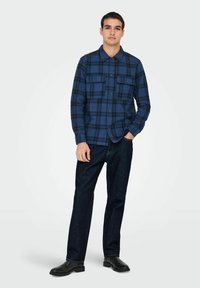 Chemise en flanelle à carreaux bleus avec deux poches poitrine et fermeture à boutons, associée à un jean en denim foncé et des bottes noires, se tenant sur un fond neutre.