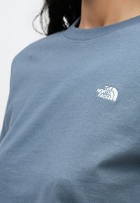 Nahaufnahme einer Person, die ein blaues Sweatshirt mit Rundhalsausschnitt trägt, auf dessen Brust ein kleines weißes The North Face-Logo zu sehen ist.