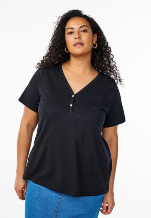 Kobieta o kręconych włosach, nosząca złote kolczyki-koła, czarną bluzkę z krótkim rękawem i dekoltem w serek zapinaną na guziki oraz niebieską spódnicę dżinsową, stoi na tle białego tła.