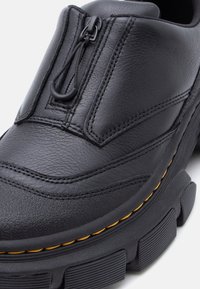 Μαύρο δερμάτινο παπούτσι με μπροστινό φερμουάρ, υφή στη μύτη, κίτρινη ραφή και χοντρή λαστιχένια σόλα με βαθιές αυλακώσεις.