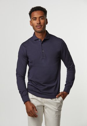 Uomo con capelli corti e ricci che indossa una polo blu navy a maniche lunghe e pantaloni beige, in piedi con una mano in tasca.