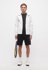 Νέος άνδρας στέκεται κρατώντας ρακέτα τένις, φοράει λευκή ζακέτα με φερμουάρ, μαύρες σόρτς, μαύρο μπλουζάκι, λευκές κάλτσες και λευκά αθλητικά παπούτσια.