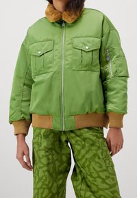 Grön bomberjacka med brun pälskrage, stora bröstfickor och ribbad mudd. Materialet har en slät, glänsande yta.
