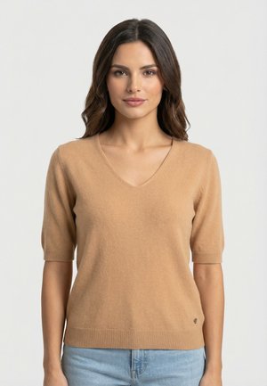 Femme aux cheveux bruns mi-longs, portant un pull beige à manches courtes et col en V, ainsi qu'un jean bleu clair, debout devant un fond uni.