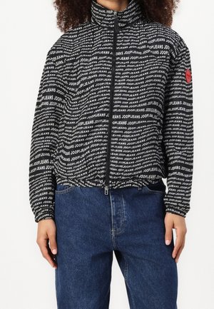 Osoba ubrana w czarną kurtkę zapinaną na zamek, pokrytą powtarzającym się białym napisem "JOOP! JEANS" oraz niebieskie dżinsy, na białym tle.