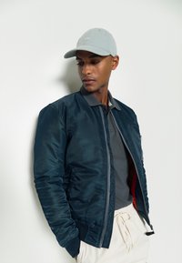 Giacca bomber navy con una texture liscia e chiusura a zip, abbinata a un polo grigio. Il modello indossa un cappellino azzurro chiaro, posando su uno sfondo bianco.