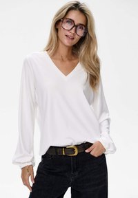 Weißes Langarmoberteil mit V-Ausschnitt und gerafften Manschetten. Kombiniert mit dunklen Jeans und einem braunen Gürtel mit einer goldenen Schnalle.