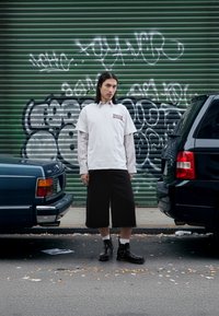 T-shirt blanc porté sur un haut à manches longues rayé, associé à un short noir jusqu'aux genoux et des bottines noires. Fond vert couvert de graffitis.