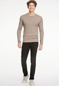 Ljusbrun tröja med långärmad ribbad struktur, kombinerad med svarta smala jeans och beige sneakers, på en modell med lockigt hår.